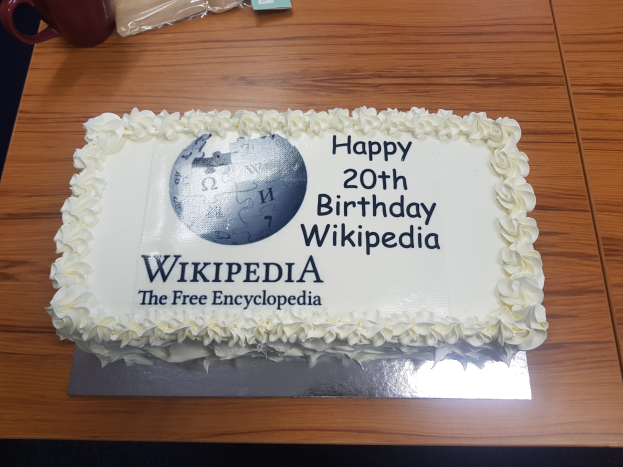 Geburtstagstorte mit "Happy 20th Birthday Wikipedia" darauf, auf einem Tisch mit einer Tasse, Hülle und anderen Gegenständen.