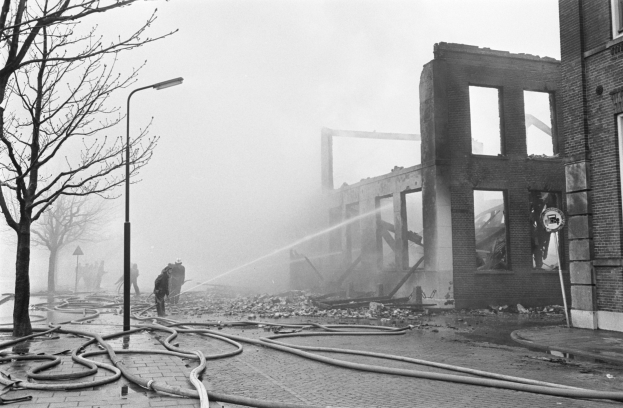 Schwarz-weiß-Bild eines feuerbeschädigten Gebäudes mit Feuerwehreinsatz, umgeben von Trümmern, Menschen, Rohren, einer Straßenlaterne, einem Schild, Bäumen und einem bewölkten Himmel.