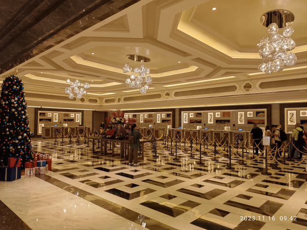 Eine große Lobby mit einem zentralen Weihnachtsbaum, umgeben von Geschenkboxen und festlicher Deko, mit Menschen, Absperrböcken, Blumentischen, gerahmten Fotos an den Wänden und beleuchteter Decke, die eine gemütliche Atmosphäre schaffen.