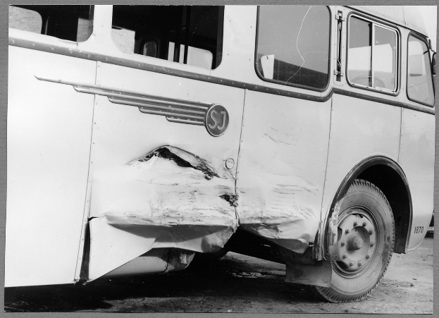 Schwarzes und weißes Foto eines auf der Straße liegenden Busses mit eingedrückter Front, zersplitterten Fenstern und offener Fahrertür, die den Innenraum zeigt.