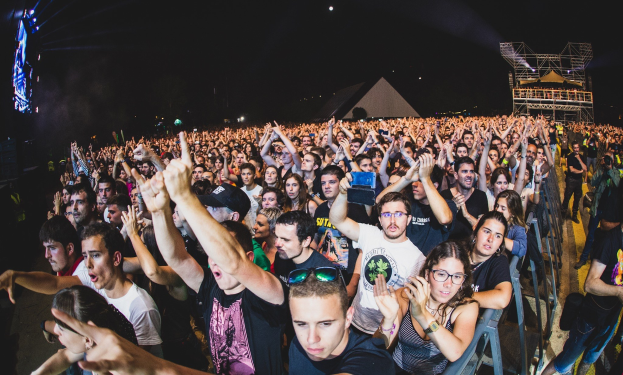 Menschenmenge auf einem Musikfestival mit erhobenen Händen, sitzende und stehende Besucher, ein Metallzaun auf der rechten Seite, Gebäude und Lichter im Hintergrund und ein dunkler Himmel.