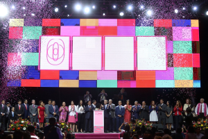 Eine Gruppe von Menschen auf einer Bühne mit einem Podium, Blumengestecken auf beiden Seiten und einem großen Bildschirm im Hintergrund, was die Eröffnungszeremonie des 2018 China International Film Festivals anzeigt.