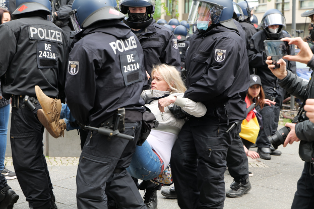 Eine Gruppe von Polizisten in Einsatzausrüstung eskortiert eine Frau in einem weißen und blauen Kleid, wobei ein Beamter ihren Arm hält, während eine Person rechts fotografiert; Gebäude, Bäume und Himmel sind im Hintergrund sichtbar.