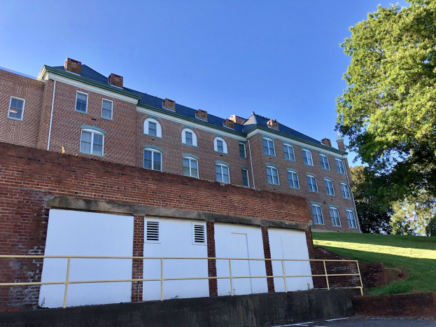 Großes Backsteingebäude mit zahlreichen Fenstern, umgeben von einem Metallzaun, auf dem Campus der University of Virginia, umgeben von einem Weg, Gras, Bäumen und einem bewölkten Himmel.