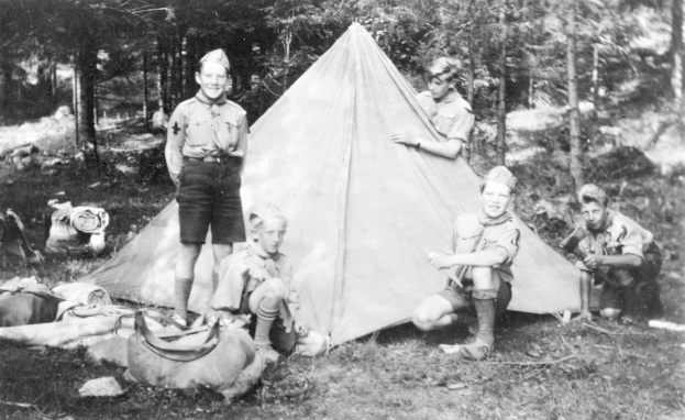 Schwarz-weiß-Foto von Pfadfindern in Uniformen, die sich um ein Zelt in einem bewaldeten Gebiet versammelt haben, einige sitzen auf dem Boden, während andere stehen, mit verstreuten Taschen und Equipment.