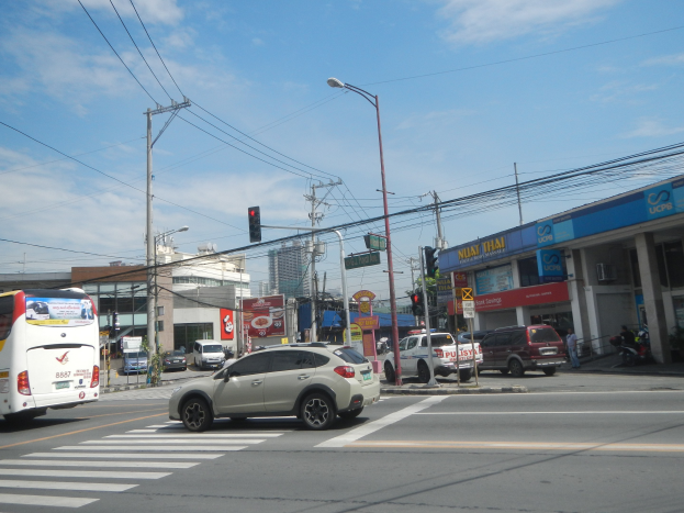 Eine Stadtstraße mit fahrenden Autos und Bussen, elektrischen und Laternenmasten mit Drähten, Verkehrsampeln, Gebäuden mit Namensschildern und einem bewölkten Himmel im Hintergrund.