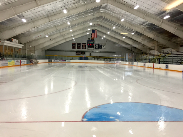 Große Indoor-Eisfläche mit Deckenbeleuchtung, Werbetafeln, einer Wand im Hintergrund, Treppen mit Geländern und einigen Menschen, was auf eine Sportanlage hinweist, die likely für Hockey verwendet wird.
