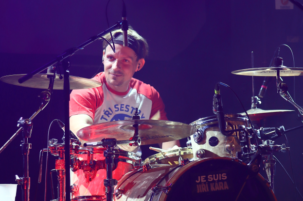 Ein Mann in einem roten und weißen T-Shirt spielt auf der Bühne Schlagzeug und lächelt umgeben von Mikrofonen.