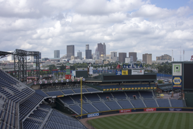 Baseballstadion mit einer Stadtkulisse im Hintergrund, gefüllt mit Stühlen, Pfählen und Brettern, auf Gras mit Bäumen und Gebäuden in der Ferne unter einem bewölkten Himmel.