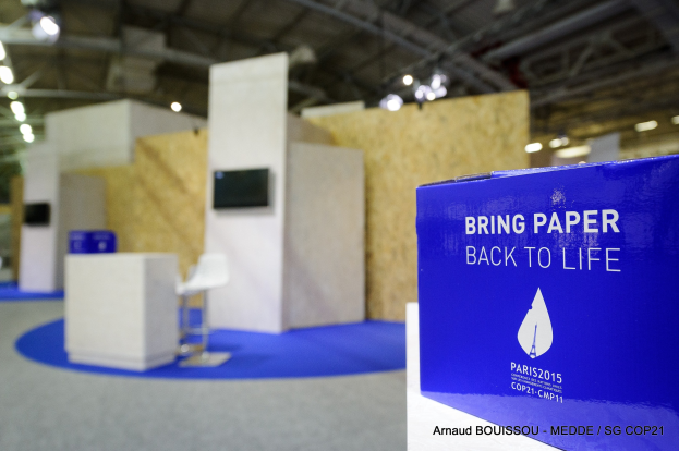 Ein Messestand mit einem blauen und weißen Schild, auf dem "Bring Paper Back to Life" steht, auf der rechten Seite, mit unscharfen Stühlen, Tischen, einem Wandfernseher und Deckenlampen im Hintergrund.