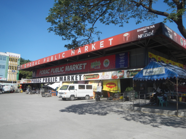 Ein ƶffentlicher Markt mit einem vor ihm geparkten Van, umgeben von Gebäuden, Geschäften, Namensschildern, Kraftfahrzeugen auf der Straße, Personen unter einem Zelt sitzend, Geländern, Bäumen und einem klaren blauen Himmel.