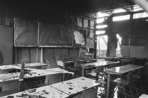 Schwarzes und weißes Foto eines Klassenzimmers mit verkohlten Wänden, zerbrochenen Fenstern und leeren Tischen und Stühlen.