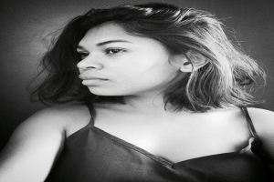 Eine selbstbewusste Frau in einem schwarzen Kleid posiert in einem Schwarz-Weiß-Foto, ihr glatter Bob und das natürliche Make-up unterstreichen ihre selbstbewusste Haltung vor einer Wand.