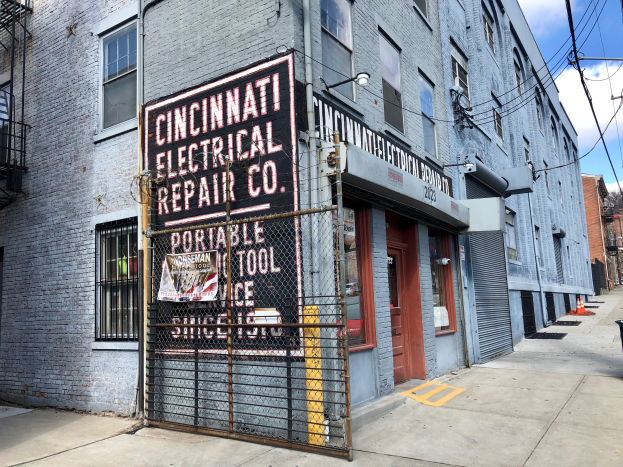 Außenansicht des Cincinnati Electrical Repair Co. Gebäudes mit einem Schild, einem Metallzaun, einem Müllcontainer, Strommasten, Drähten, Bäumen und einem bewölkten Himmel.