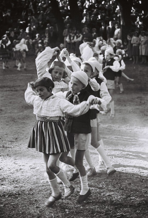 Schwarz-weiß-Foto von Kindern, die in einem Park tanzen, mit einigen Zuschauern und Bäumen im Hintergrund.