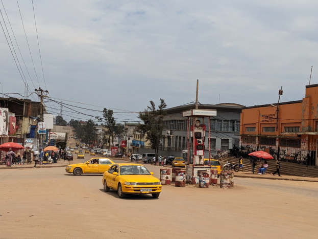 Ein gelbes Taxi fährt auf einer belebten Stadtstraße mit Gebäuden an den Seiten entlang, während Fußgänger mit Regenschirmen auf dem Gehweg darunter unter bewölktem Himmel gehen.