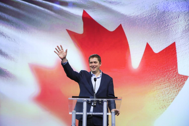 Der kanadische Premierminister Alex Salmond steht an einem Rednerpult mit Mikrofonen, lächelt und schaut in die Kamera, vor einer kanadischen Flagge und einem Bildschirm im Hintergrund.