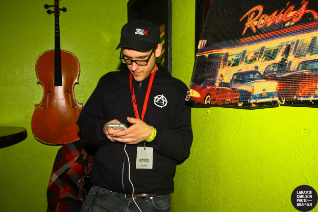 Eine Person mit einer Mütze und einem Ausweis hält ein Mobiltelefon in der Hand und schaut auf dessen Bildschirm, mit einer Gitarre links und einem Plakat rechts.