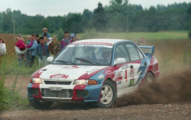 Ein Rallye-Auto, verziert mit Text und Zahlen, fährt eine Schotterstraße entlang, umgeben von einer Menschenmenge, einige halten Kameras, mit Pflanzen, Gras, Polen, Drähten, Bäumen und einem klaren blauen Himmel im Hintergrund.