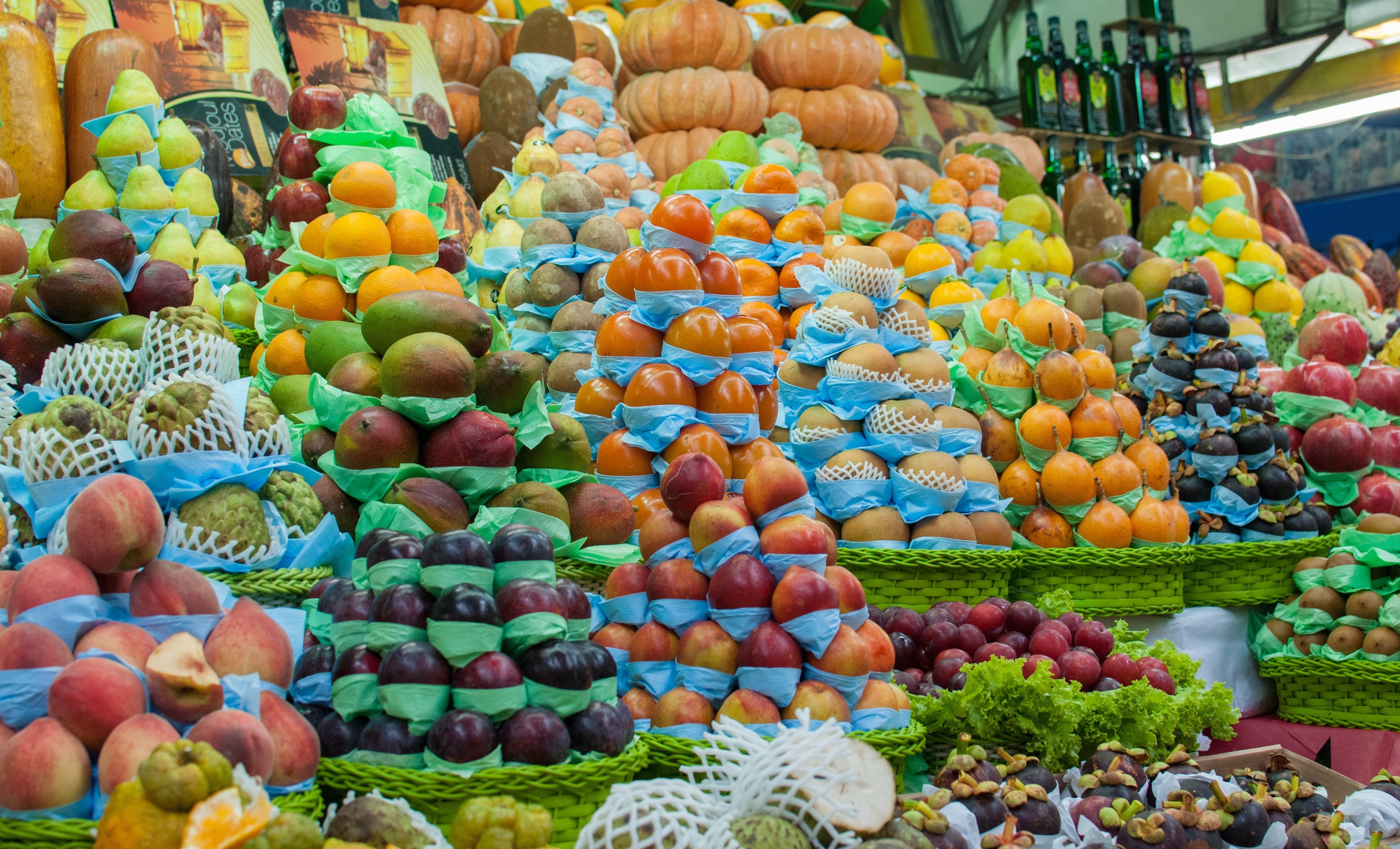 Ein Marktstand mit Äpfeln, Orangen, Birnen und anderen Früchten und Gemüsen, mit Flaschen auf Regalen und Deckenbeleuchtung.