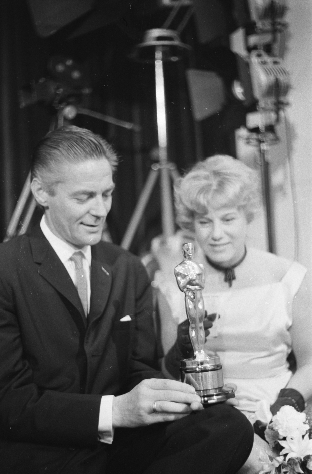 Ein Mann in einem schwarzen Blazer, weißem Hemd und Krawatte hält eine Oscar-Statue in der Hand und sitzt neben einer Frau, die einen Blumenstrauß hält, mit ein paar Lichtern im Hintergrund, alles in Schwarz-Weiß.