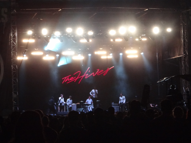 Eine Gruppe von Menschen, die auf einer Bühne bei einem Konzert spielen, wobei einige Instrumente halten und einer in ein Mikrofon singt, während das Publikum unten zusieht.