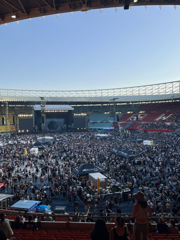 Eine große Menschenmenge sitzt und steht in einem Stadion mit Geländern, Zelten und Beleuchtung unter einem überdachten Bereich mit Lichtern, was auf ein Konzert in der O2 Arena in London hindeutet.