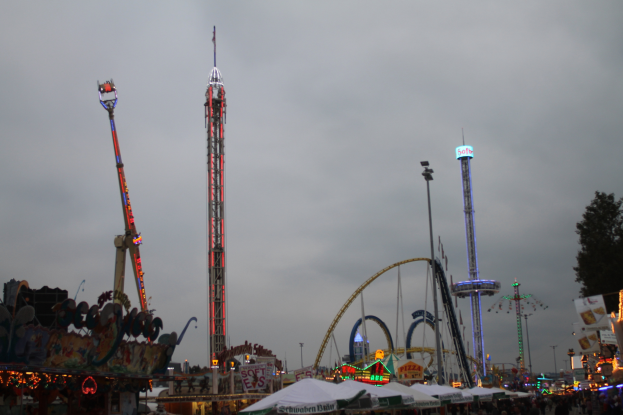 Eine nächtliche KarnevalsSzene mit einer Menge Menschen, die sich um beleuchtete Fahrgeschäfte, Pfähle und Bäume versammelt haben, unter einem bewölkten Himmel.