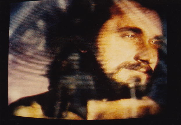 Ein Mann mit langem dunklem Bart und Haar, der ein weißes Hemd trägt, erscheint auf einem Fernseher mit dunklem Hintergrund und schaut direkt in die Kamera.