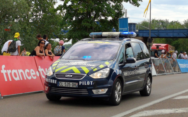 Polizeiauto fahrt auf einer Stra├če neben einer Menschenmenge, mit einer Banner auf der linken Seite des Autos und Gel├ädern mit Bannern dahinter; B├Ąume, eine Brücke, eine Flagge und ein bew├Âlktes Himmel sind im Hintergrund sichtbar.