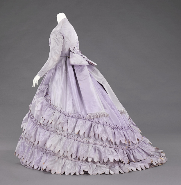 Eine Schaufensterpuppe in einem lila Ballkleid mit engem Oberteil, vollem bodenlangen Rock und Rüschen an den langen Ärmeln vor einem grauen Hintergrund.