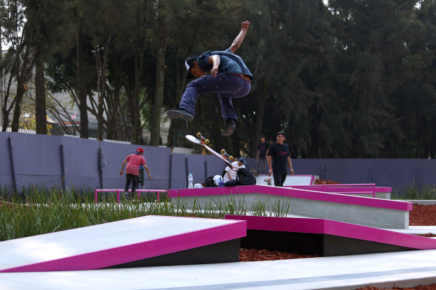 Ein Skateboarder zeigt einen Sprung auf einer Rampe, umgeben von Zuschauern mit Skateboards und verstreuten Gegenständen, vor einer Kulisse aus Bäumen, Gebäuden und einem klaren Himmel.