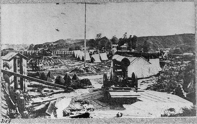 Schwarz-weiß-Foto einer Baustelle mit einer Maschine im Vordergrund, umgeben von Baracken, Bäumen, Hügeln und einem Himmel im Hintergrund, mit Text unten lesend "Die Bauarbeiten an einer neuen Brücke in der Stadt San Francisco, Kalifornien."