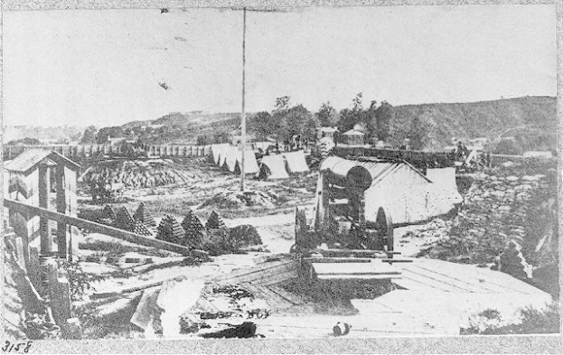 Schwarz-weiß-Foto einer Baustelle mit einer Maschine im Vordergrund, umgeben von Baracken, Bäumen, Hügeln und einem Himmel im Hintergrund, mit Text unten lesend "Die Bauarbeiten an einer neuen Brücke in der Stadt San Francisco, Kalifornien."