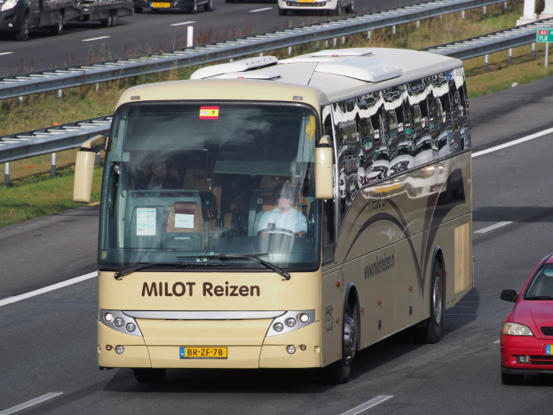 Ein Minibus mit sichtbaren Insassen fährt neben einem roten Auto auf einer von Geländern und Gras gesäumten Autobahn, mit textbehafteten Säulen und Tafeln im Hintergrund.