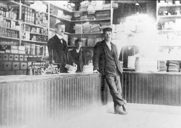 Schwarzes und weißes Foto von drei Männern an einer Ladentheke mit Gegenständen, vor einem Regal mit Büchern und Artikeln während eines scheinbaren Raubüberfalls.