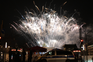 Polizeiauto vor einer Menschenmenge während einer Silvesterfeier in Berlin geparkt, mit farbenfrohen Feuerwerken, die den Himmel und die Gebäude erhellen.