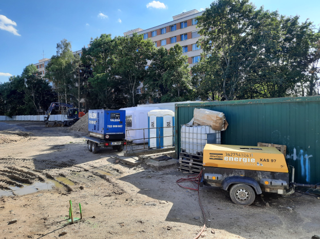 Baustelle mit einem geparkten Lkw, Containern und einem Fahrzeug vor einem Gebäude, umgeben von Bäumen, Pfählen und anderen Gegenständen unter einem bewölkten Himmel.