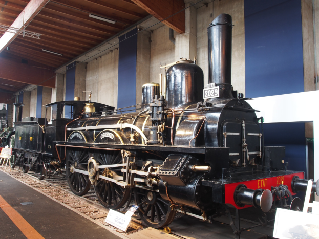 Alte Dampflokomotive in einem Museum mit umliegenden Schienen, Informationsschildern und Beleuchtungsk├Ârpern.