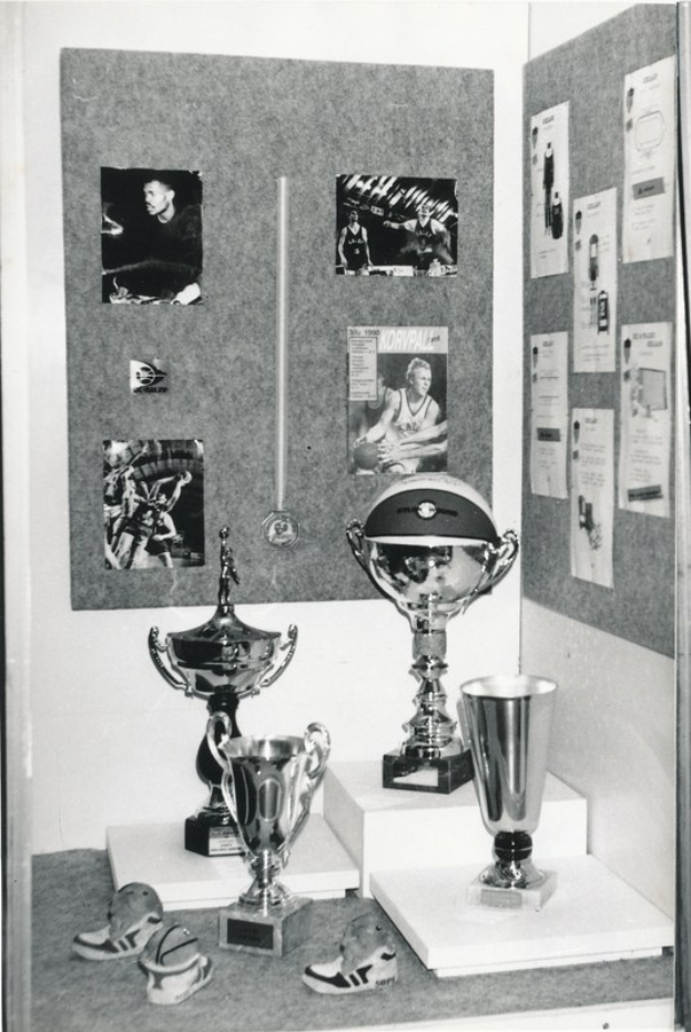 Schwarzes und wei├čes Foto von Trophen in einem Raum mit einer Tafel mit Fotos und Papieren im Hintergrund und Schuhen, die am Boden verstreut sind.