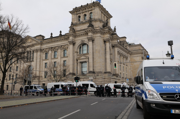 Eine Gruppe von Polizisten steht vor dem Reichstaggebäude in Berlin, Deutschland, mit Fahrzeugen, einem Zaun, Verkehrszeichen, Laternenmasten, Bäumen und Flaggen im Hintergrund, unter einem klaren Himmel.