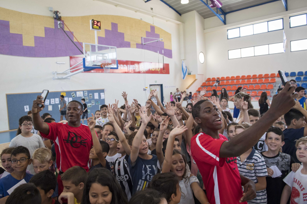 Kinder auf einem Basketballfeld mit Handys stehend, mit einer Pinnwand, Uhr, Torstange, Basketballkorb, Deckenleuchten, Stühlen und Fenstern im Hintergrund.