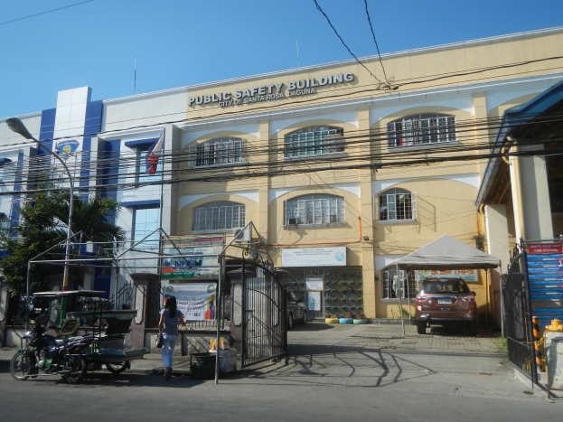 Polizeigebäude umgeben von Bäumen, Straßeninfrastruktur, Fahrzeugen, Fußgängern und einem klaren Himmel.