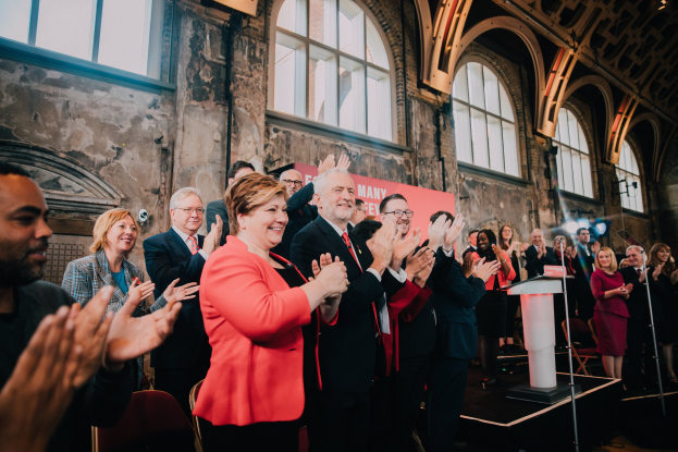 Eine Gruppe von Menschen steht vor einem Publikum, klatscht feierlich vor einem Podium mit Mikrofon, einem Banner mit der Aufschrift "Covid-19 New York City Mayor Bill de Blasio", Stühlen, einer Wand und Fenstern im Hintergrund.
