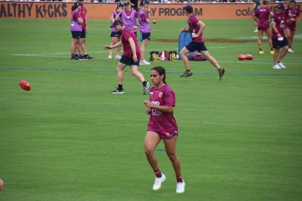 Frauen spielen Australian Rules Football auf einem grünen Feld mit verstreuten Bällen und einem Banner im Hintergrund, einige tragen Mützen und Turnschuhe.