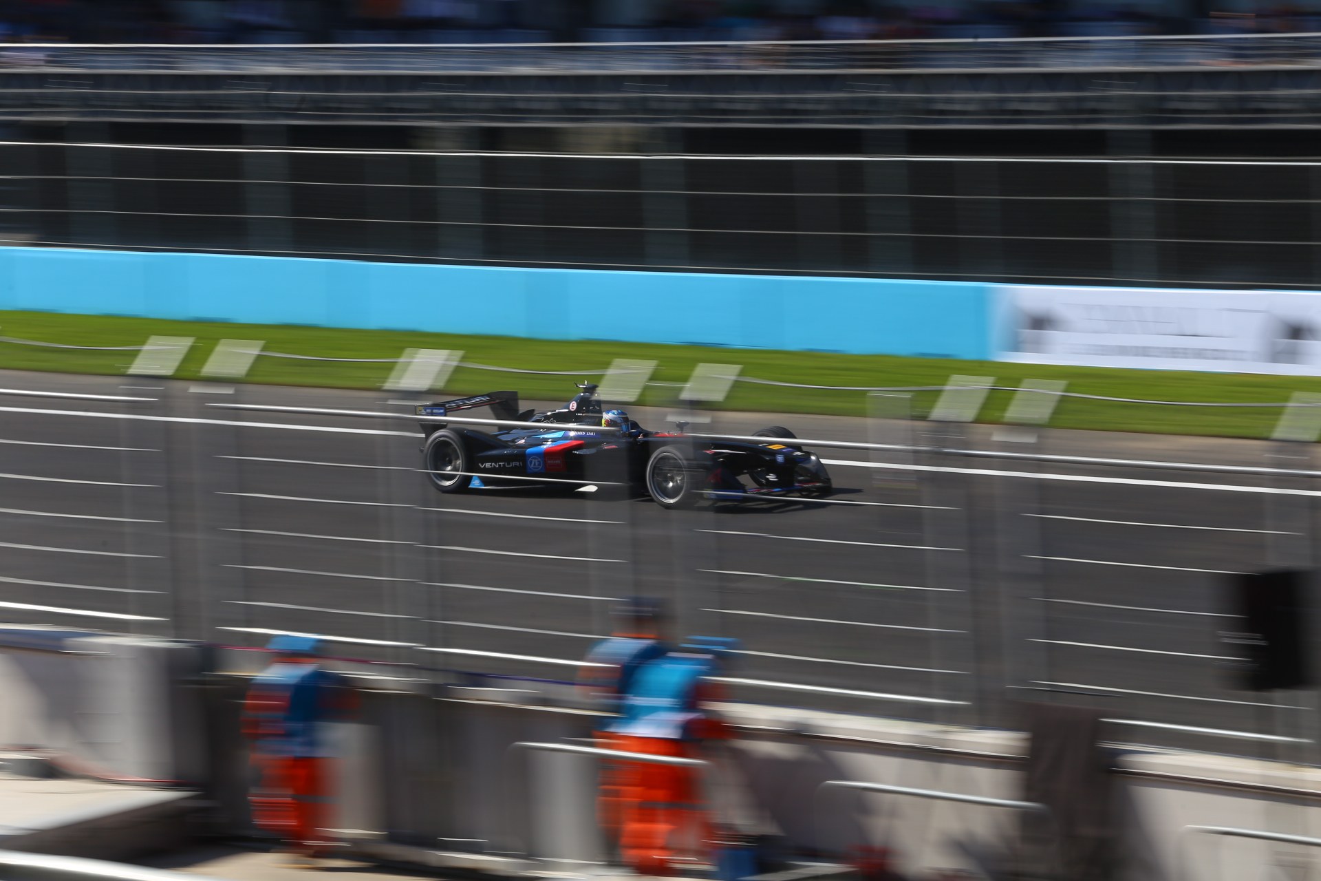 Ein fokussiertes Formel-Auto, das auf einer Rennstrecke fährt, mit Zuschauern im Hintergrund, Gras auf dem Boden und Texttafeln entlang der Strecke.