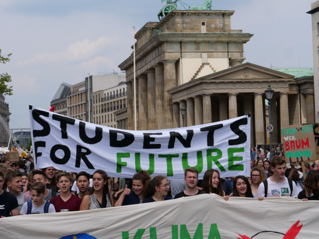 Gruppe von Schülern marschiert in Berlin mit buntem "Students for Future"-Schild an Gebäuden, Bäumen und Himmel.