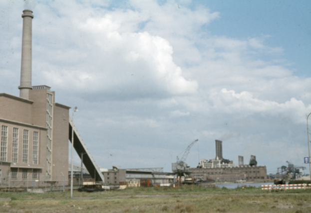 Ein altes Foto einer Fabrik mit Rauch, der aus ihren Schornsteinen aufsteigt, umgeben von verschiedenen Gebäuden, Kränen, Polen, Lampen und Schildern.