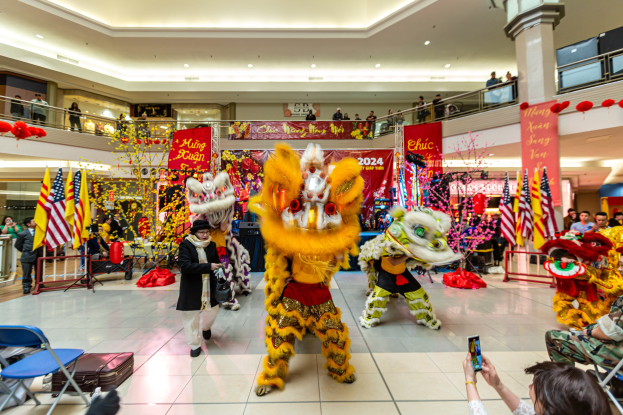 Ein lebendiges chinesisches Neujahrsfest in einem Einkaufszentrum mit Menschen in Kostümen, die tanzen, anderen, die auf Stühlen sitzen und Handys halten, sowie Flaggen, Bannern, Dekorationen, Geländern, Säulen und Deckenleuchten im Hintergrund.