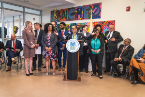 Gruppe von Menschen vor einem Podium mit Mikrofon und einer Tafel mit der Aufschrift "Sancho City Councilman Joins City Council", mit sitzenden und stehenden Teilnehmern, Flaggen, Feuerlöscher und Deckenbeleuchtung im Hintergrund.